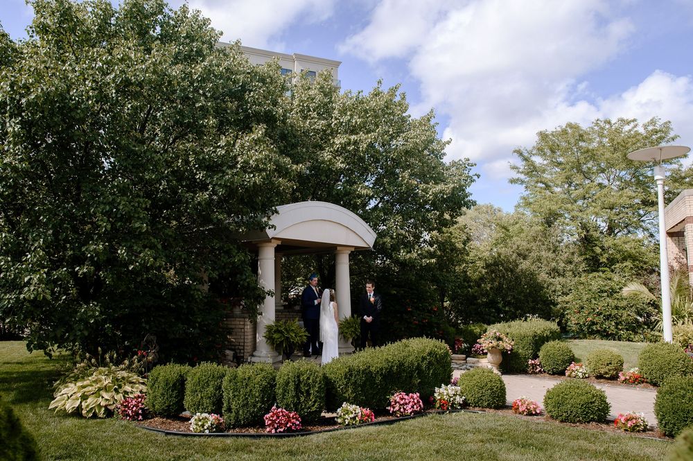White gazebo surrounded by manicured gardens with pink flowers and green trees under a partly cloudy sky.