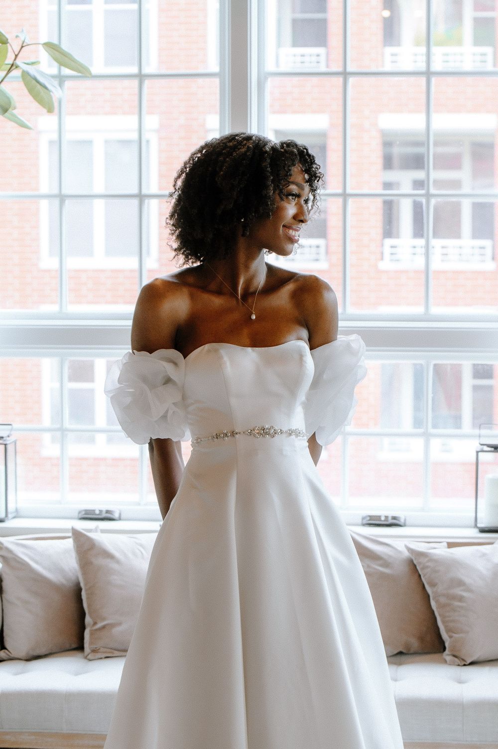 A figure in an elegant white off-shoulder wedding gown strikes different poses near sunlit windows.