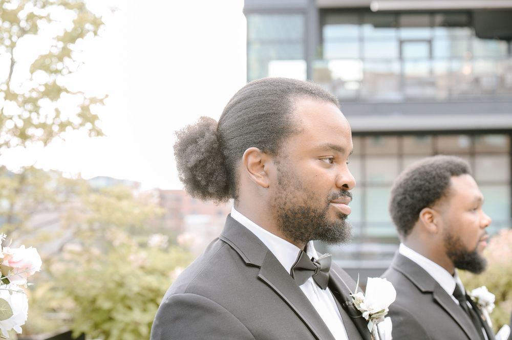 Two groomsmen wearing black tuxedos with white boutonnieres stand together outdoors on a sunny day.