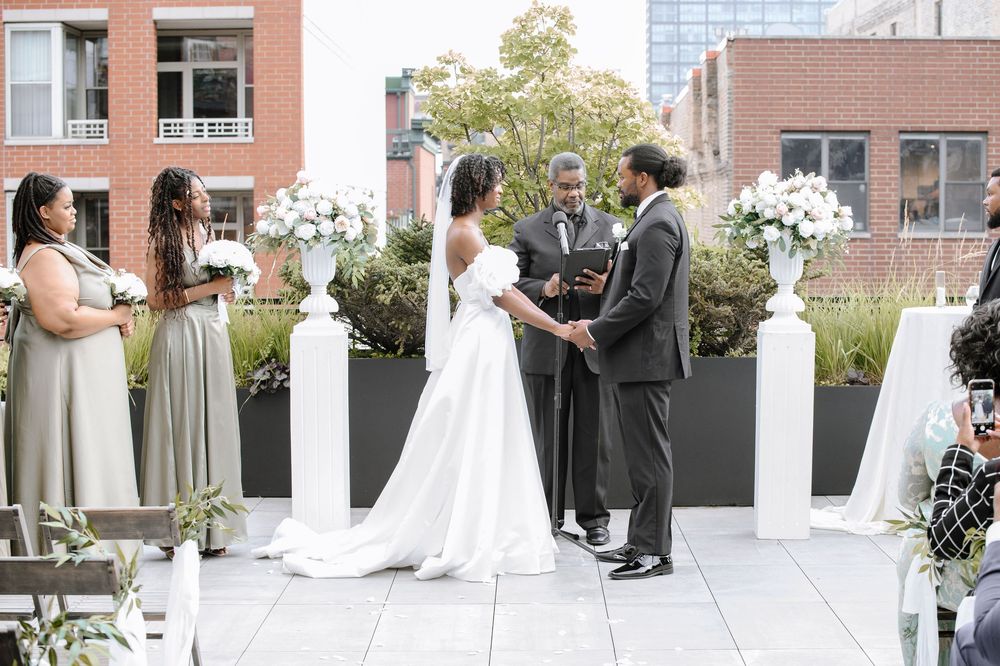 Wedding couple holds hands during intimate rooftop ceremony surrounded by bridesmaids and white floral pedestals.