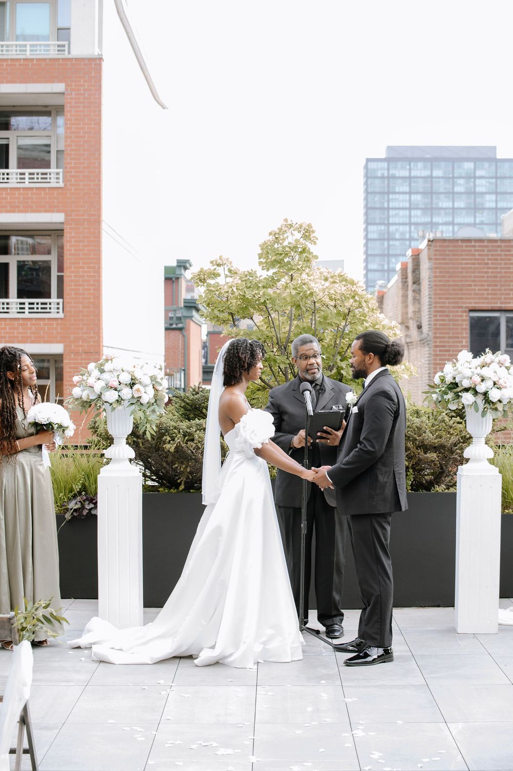 A wedding ceremony takes place on an urban rooftop terrace with floral arrangements and brick buildings in the background.
