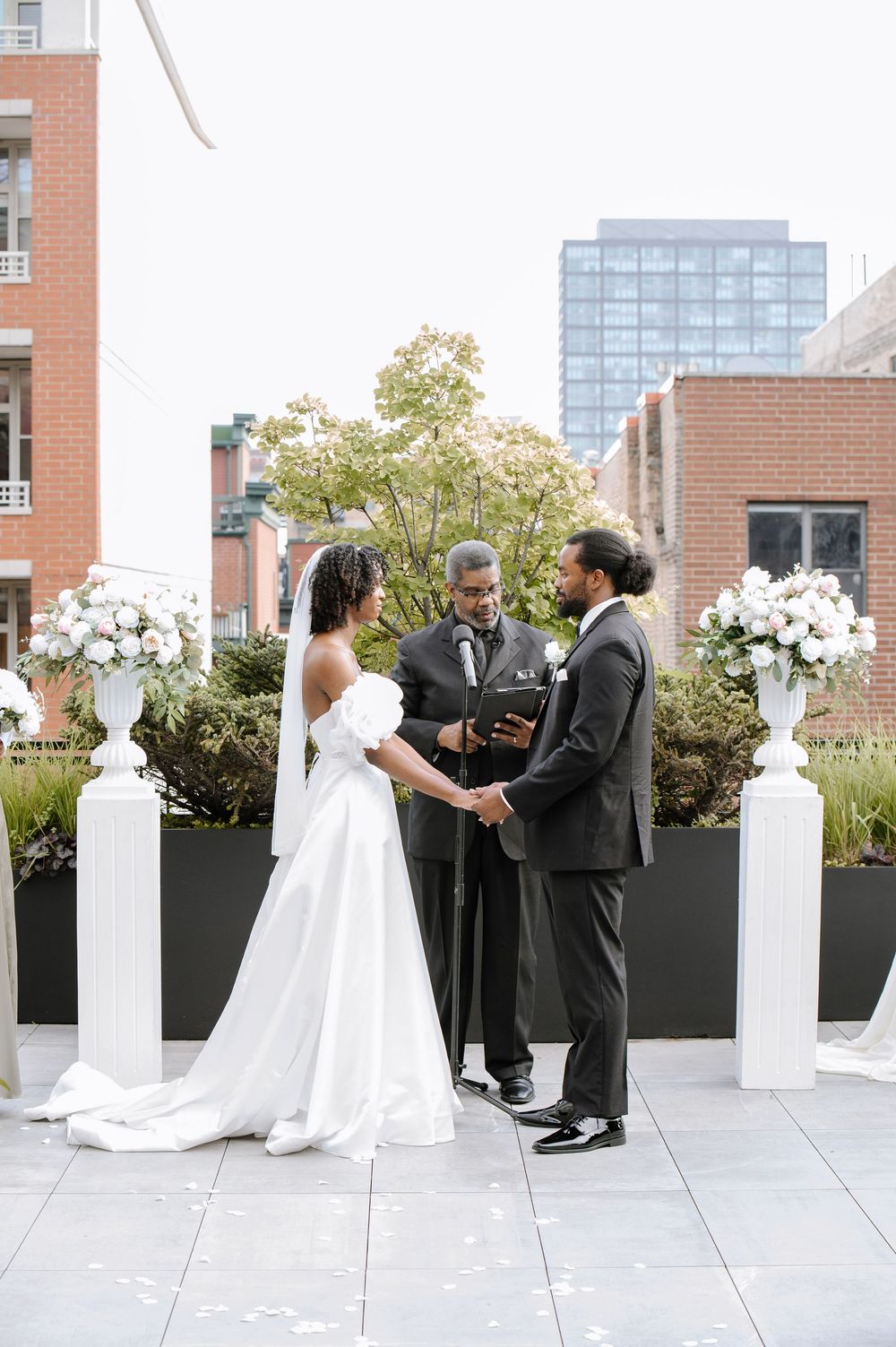 Couple in wedding attire at an outdoor ceremony against city backdrop with black planters and ornate pillars.