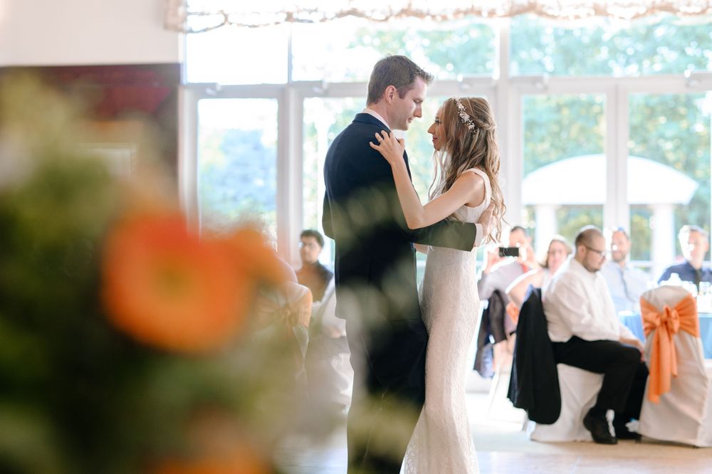 Romantic couple dancing together at a light-filled wedding reception venue with orange floral decorations in view.
