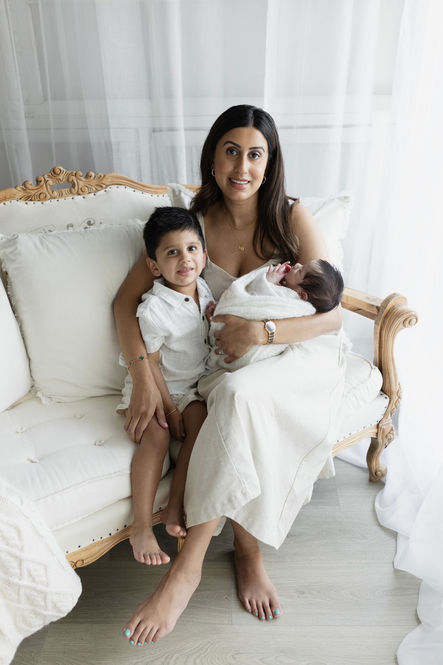 Mom sits on the all white sofa and curtain setup with her son and newborn baby boy. Elizabeth Lang Photography Surrey.
