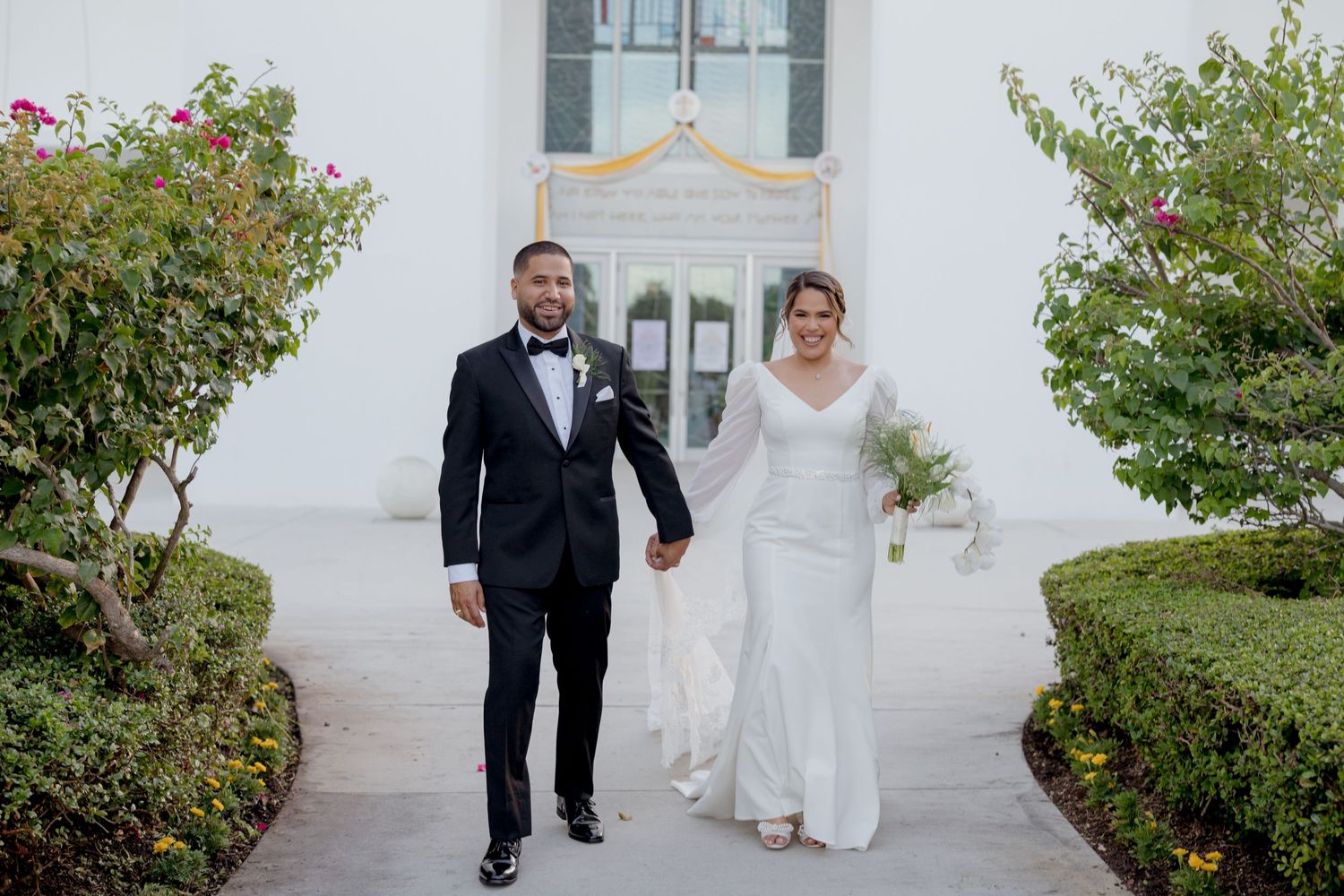 Young ring bearer walks down outdoor garden path at elegant wedding ceremony in formal attire.
