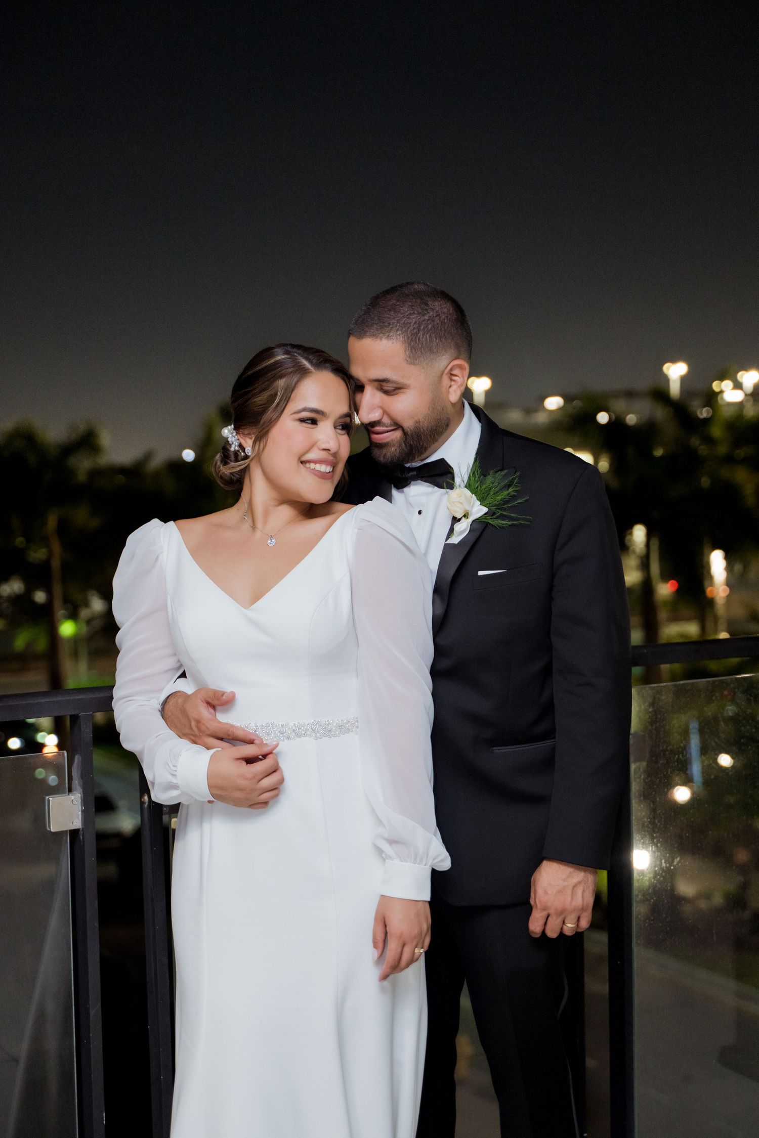 Elegant couple embraces during nighttime wedding celebration with city lights glowing in the background.