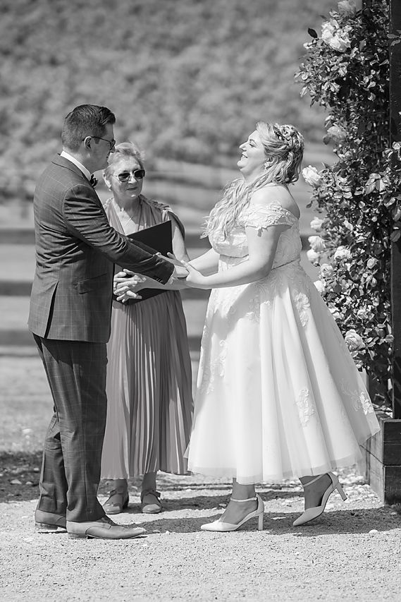 A black and white photo of a wedding ceremony taking place outdoors with a bride in a tea-length dress.