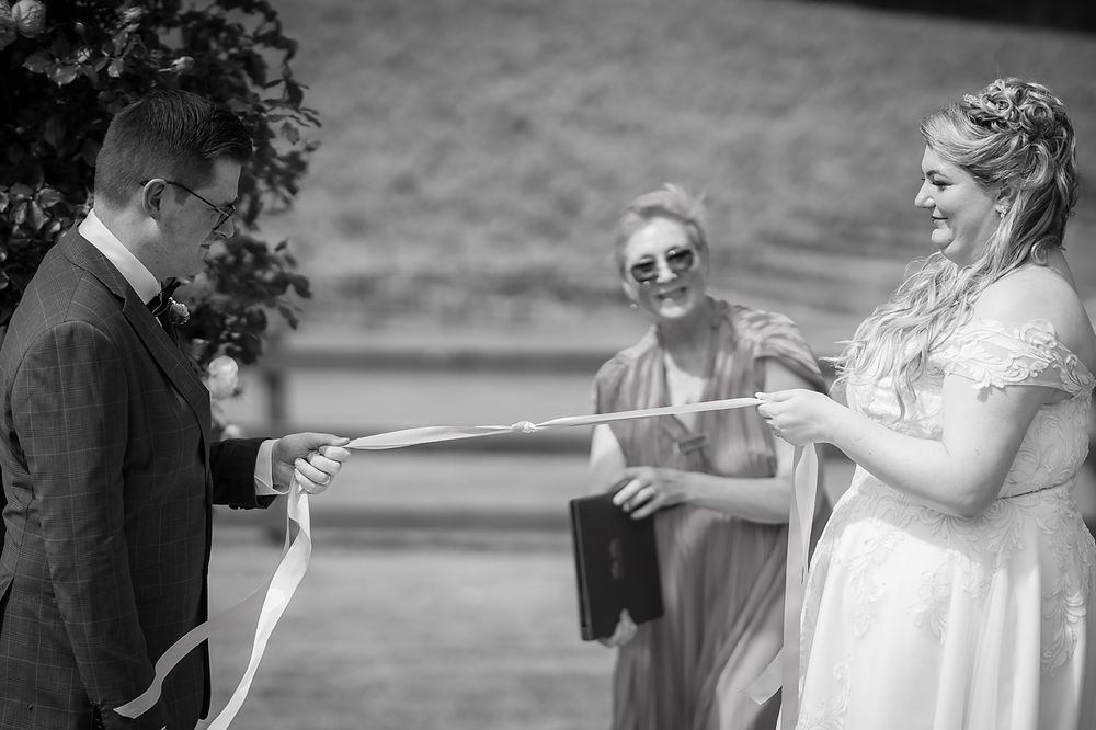 Black and white photo showing a playful moment during an outdoor wedding ceremony while exchanging ribbons.