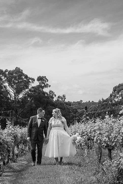 Black and white wedding photo of a couple walking hand in hand through a romantic vineyard setting.