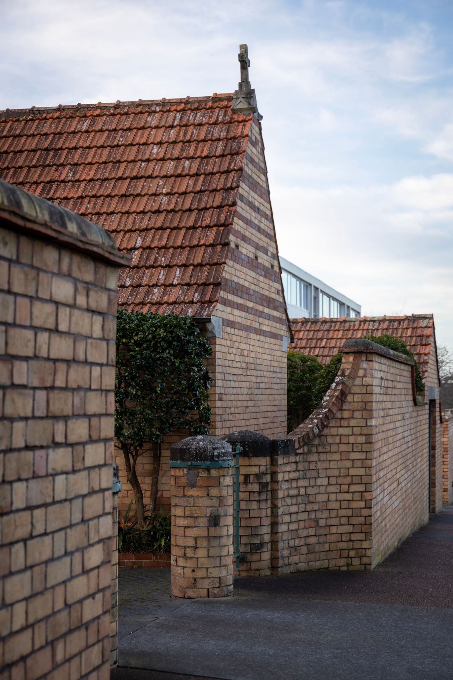 Historic brick building with red-tiled roof, arched gate and fence leading into a leafy courtyard.