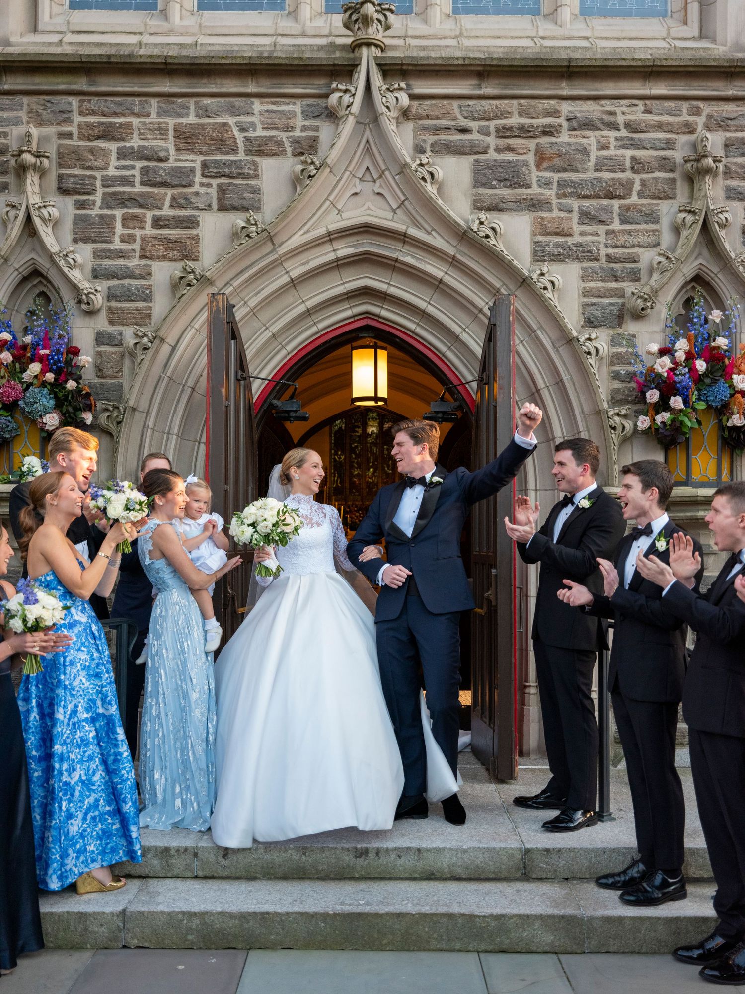 Wedding party celebrates on stone church steps as newlyweds exit through Gothic archway.