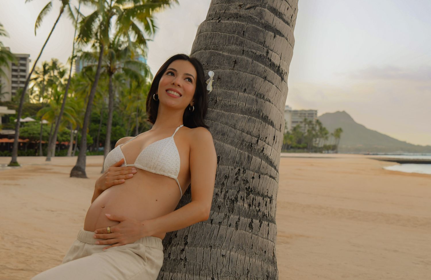 Maternity photo session at Hilton Hawaiian village, palm trees frame a scenic beach view at sunrise with Waikiki skyline in the background.