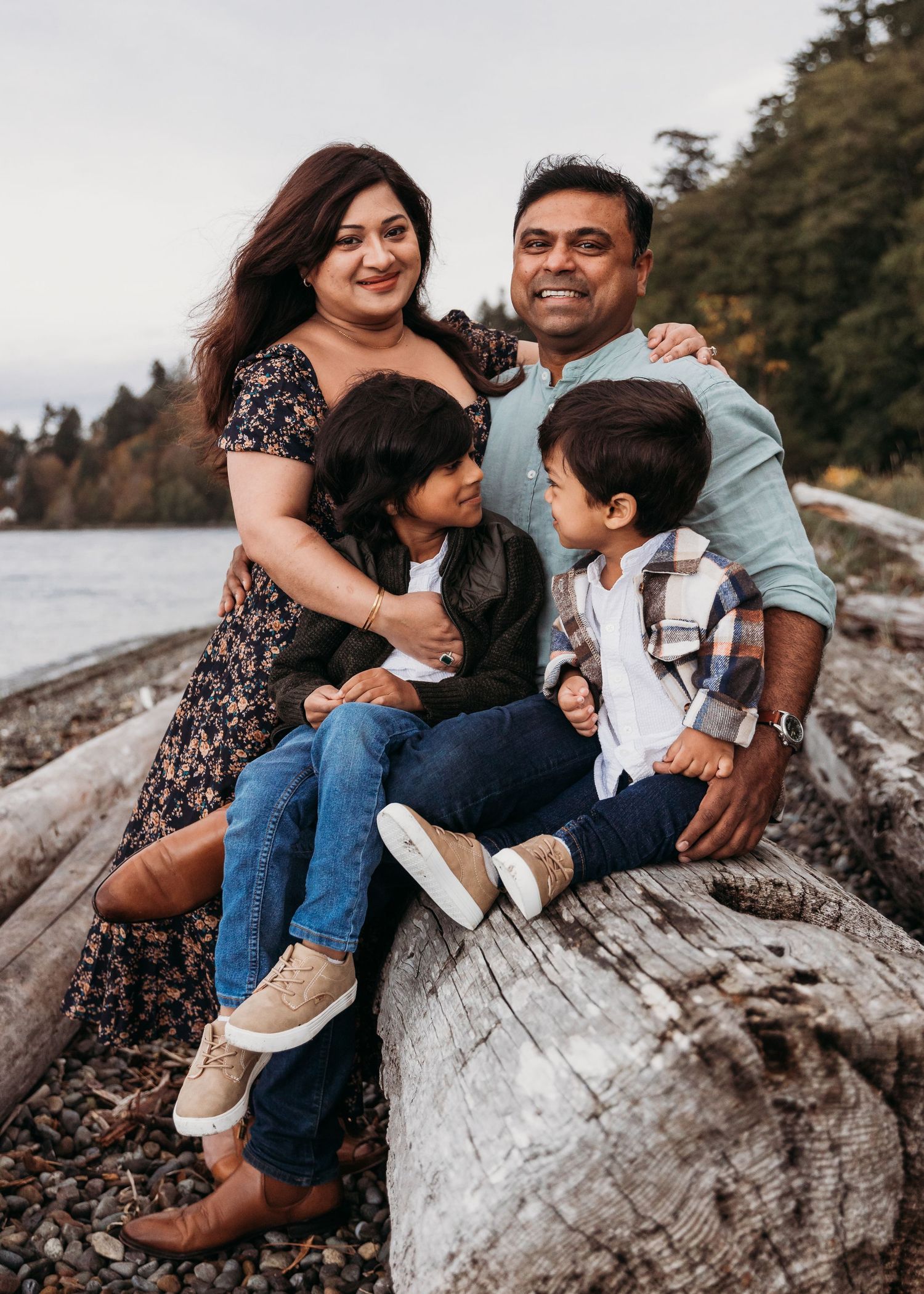 Family sits together on a driftwood log at a rocky beach during sunset, dressed casually for a photo session.