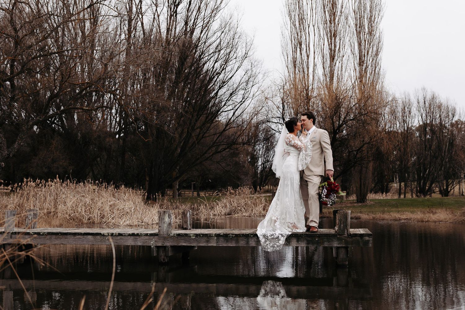 A couple in winter wedding attire stands by a lake surrounded by barren trees creating a romantic seasonal scene.