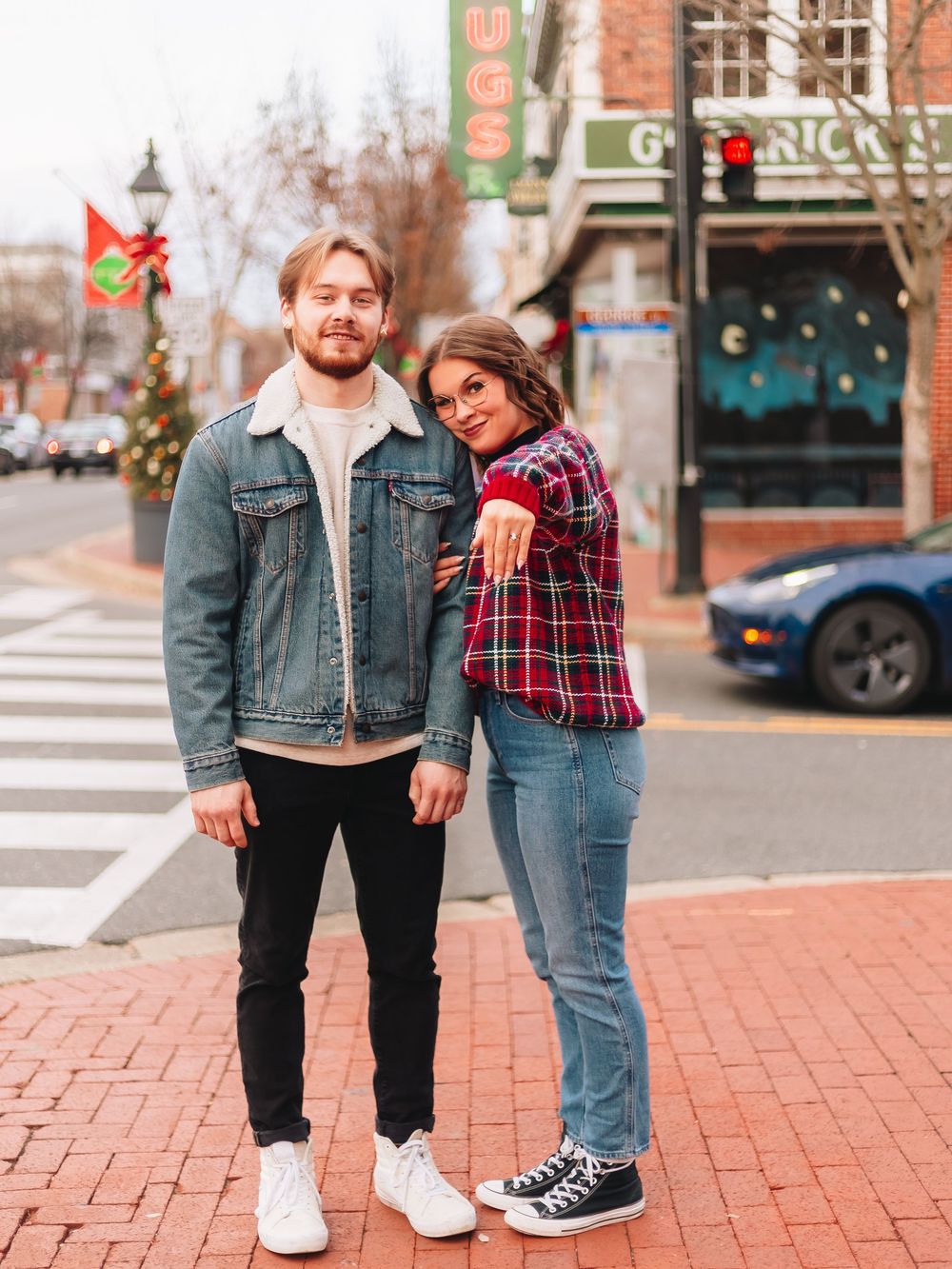 A couple poses casually on brick sidewalk in urban setting wearing denim and plaid.