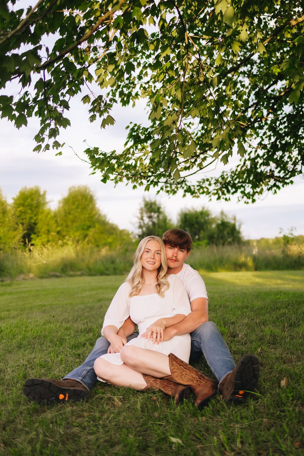 A couple relaxes under green tree branches on grassy ground during summer.