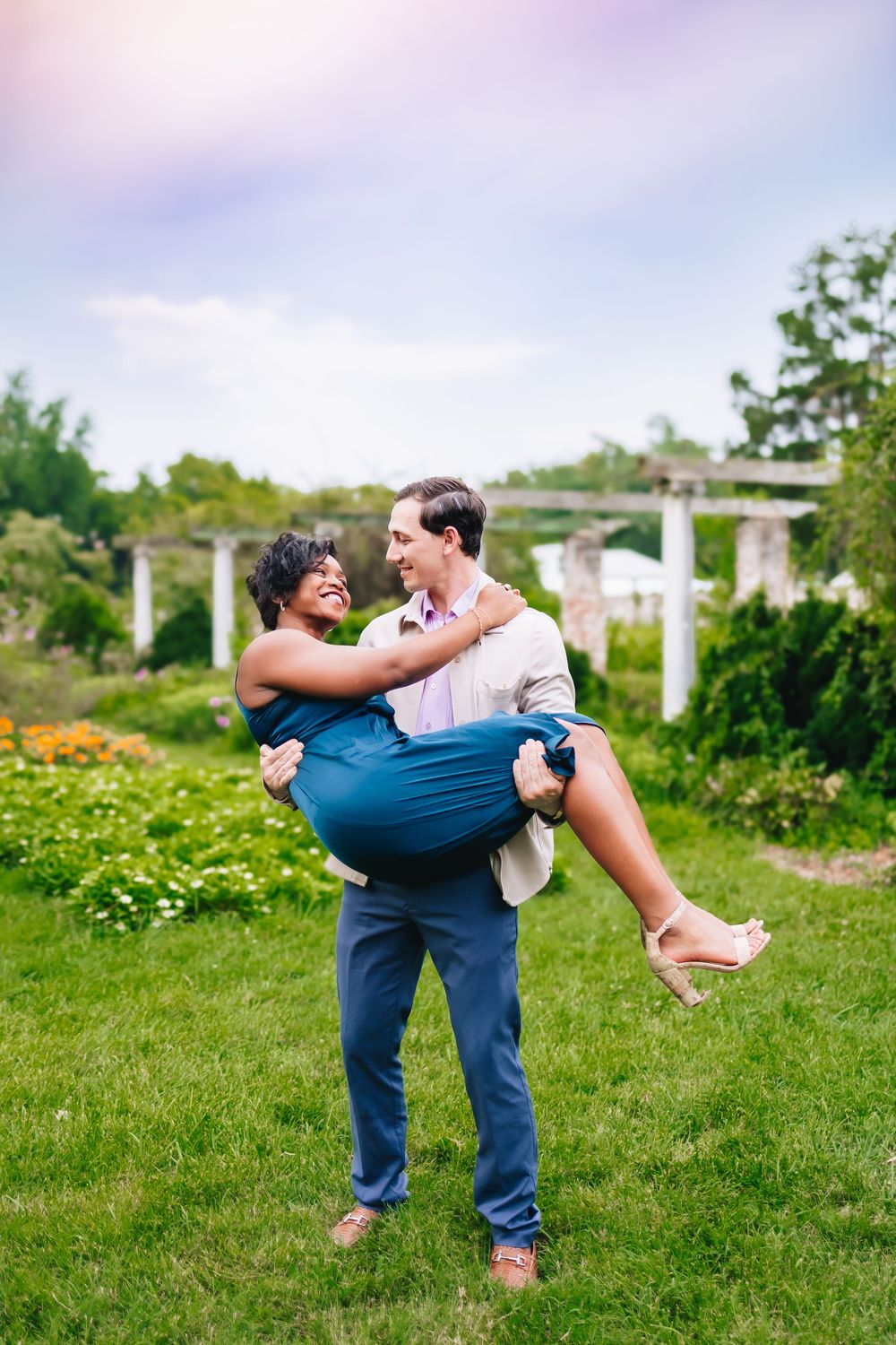 A playful lifting pose captured in a sunny garden with white pillars.