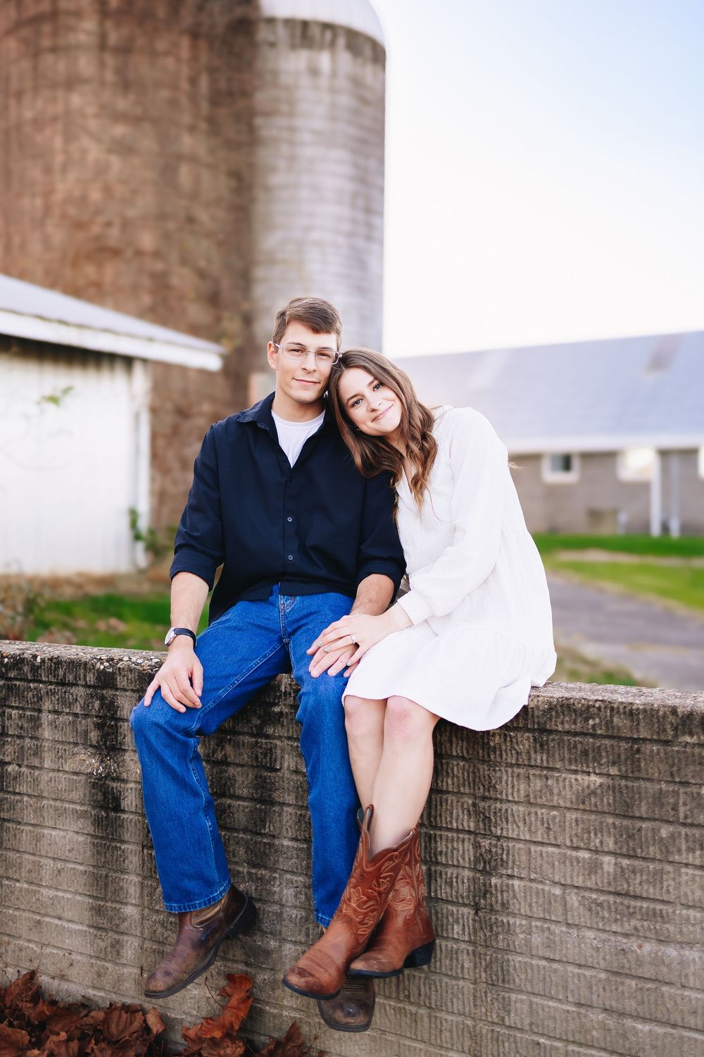 A couple sits together on a brick wall near industrial silos in casual outdoor attire.