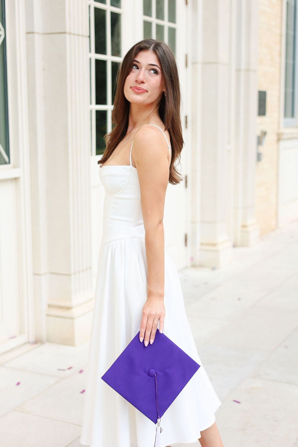 Graduate posing in white dress with purple graduation cap in front of university columns.