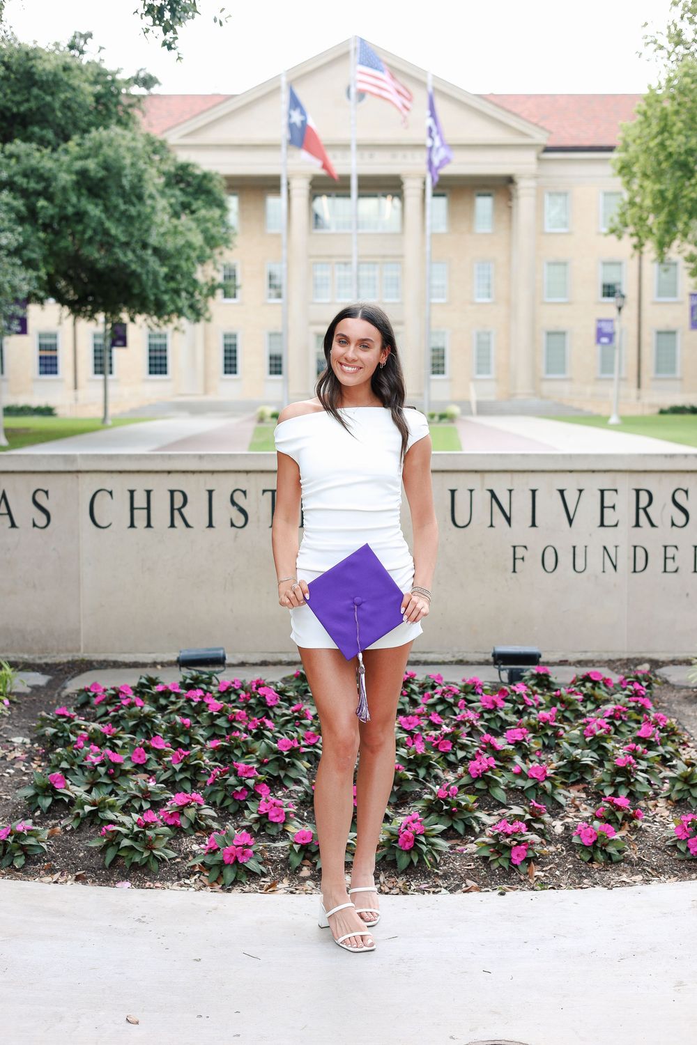 Graduate poses by university sign and pink flower garden.