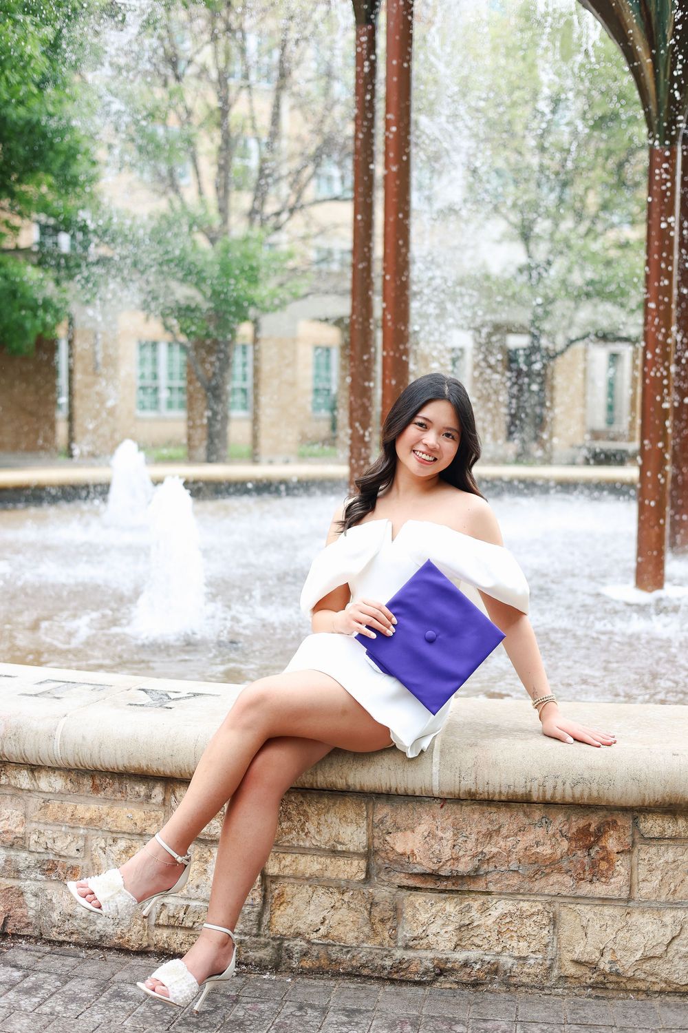 Graduate in white dress sitting casually on stone wall.