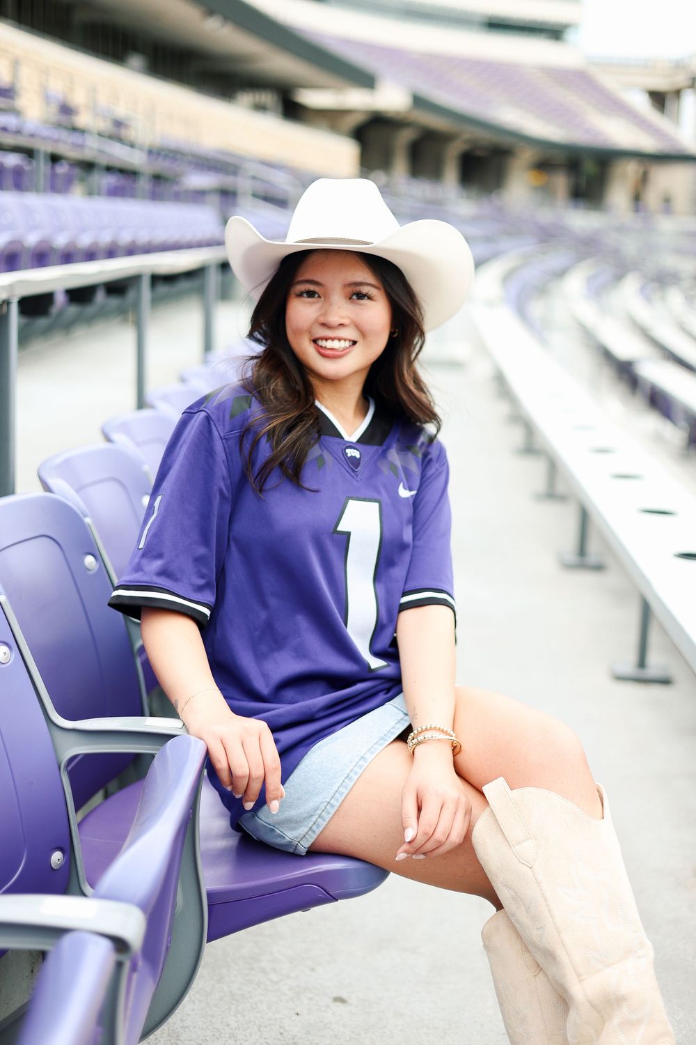 Graduate wearing cowboy hat and purple jersey sitting in stadium seats.