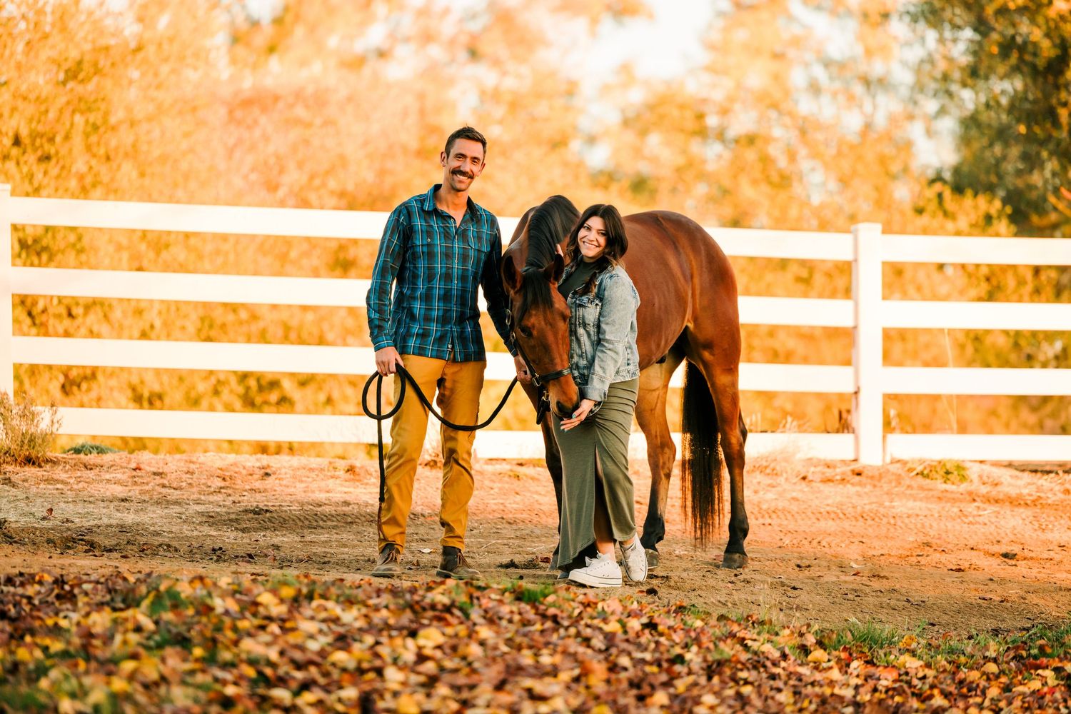 A couple and their horse enjoy an autumn sunset among fall foliage and white fences in a rustic countryside setting.