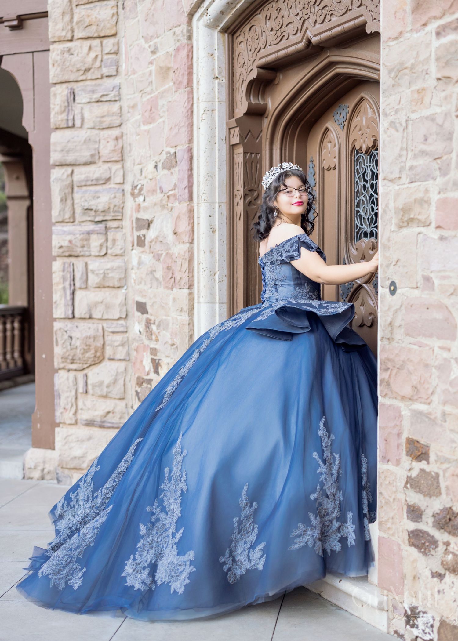 A person wearing an elegant navy blue ball gown with floral appliques poses in an ornate stone archway.