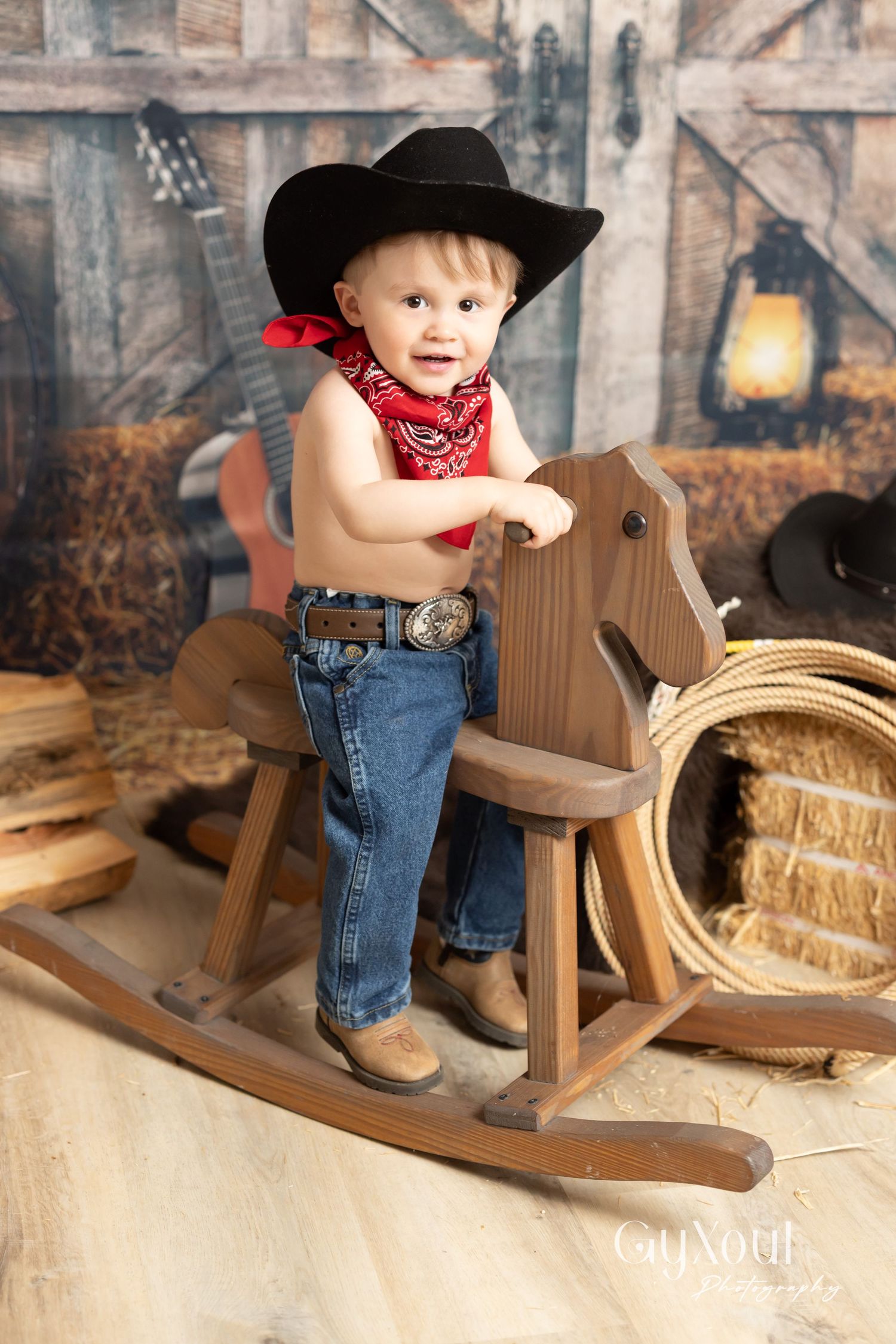 Wooden rocking horse photo with western theme featuring cowboy hat, bandana, jeans and rustic barn backdrop with lantern.