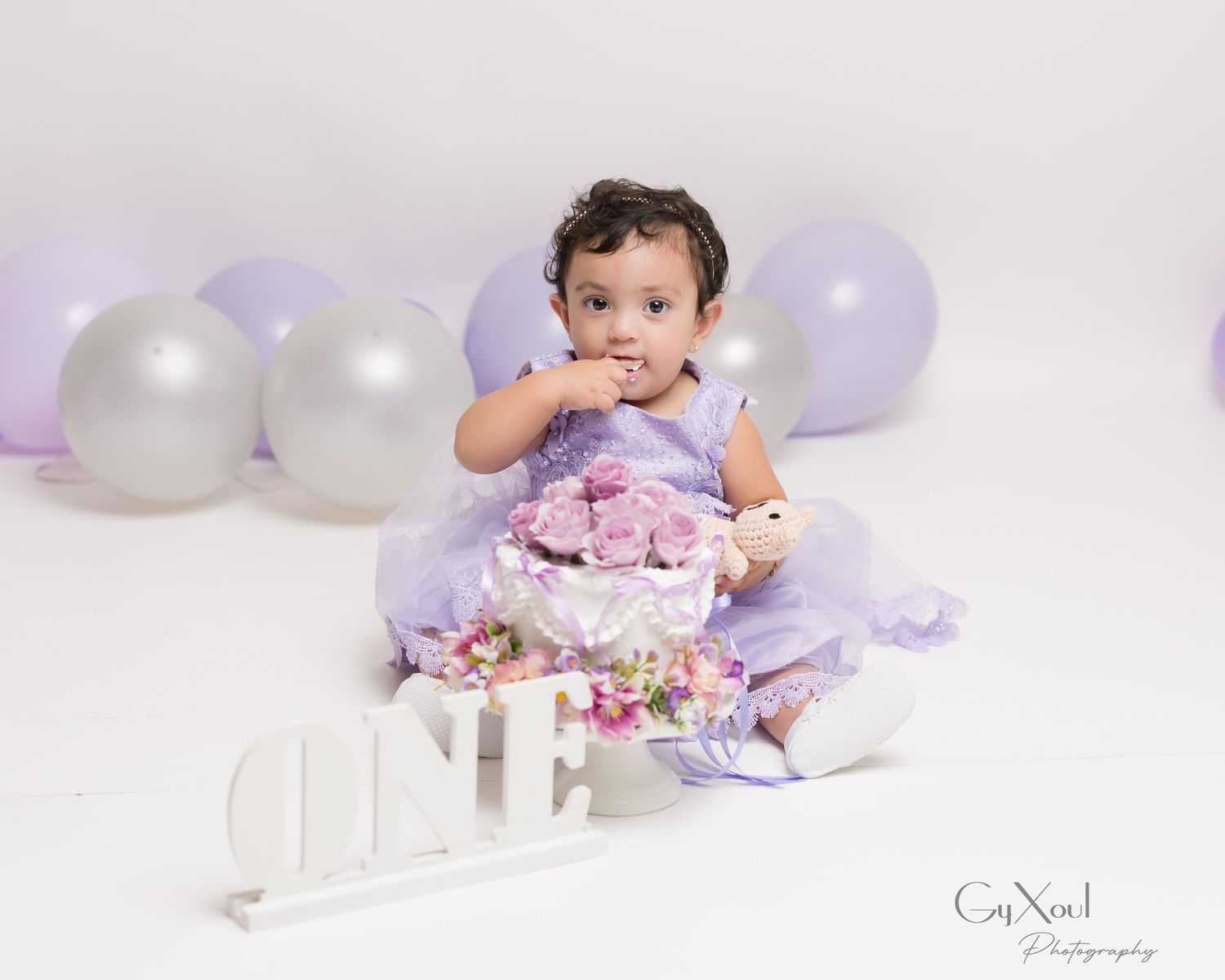 A baby in purple attire sits on the floor with cake during a first birthday photoshoot with white and purple balloons.