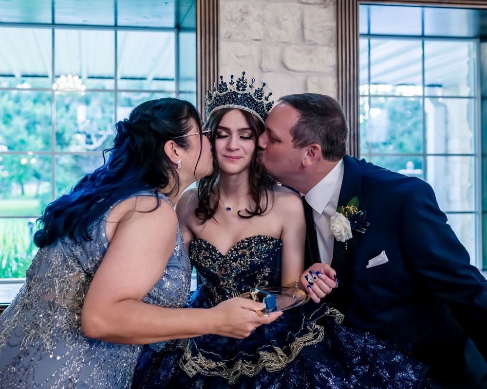 A person in a dark strapless gown and tiara receives kisses on the cheek during a formal celebration.