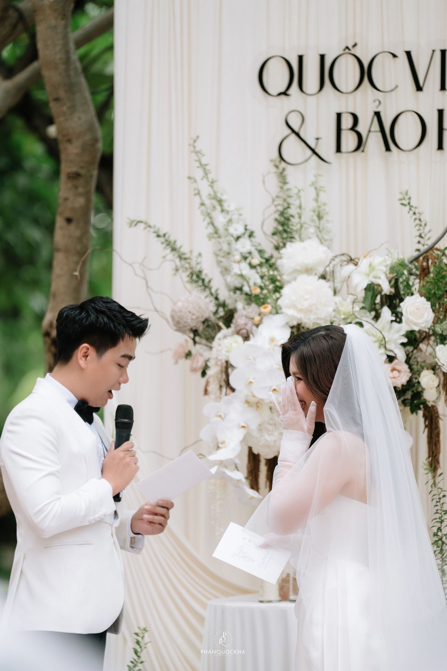 Emotional groom reads heartfelt vows to tearful bride at elegant outdoor wedding ceremony with floral backdrop.
