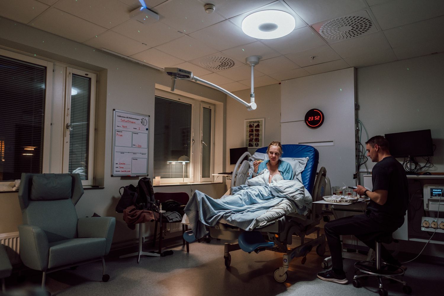 I förlossningsrummet sitter en mamma med sin bebis i förlossningssängen och äter en macka. pappan sitter på en stol bredvid sängen.
