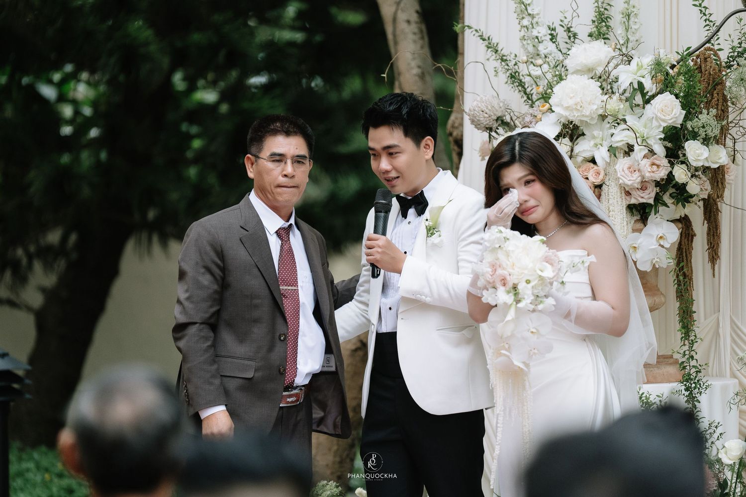 Groom speaks into microphone while emotional bride stands beside older gentleman at wedding ceremony.