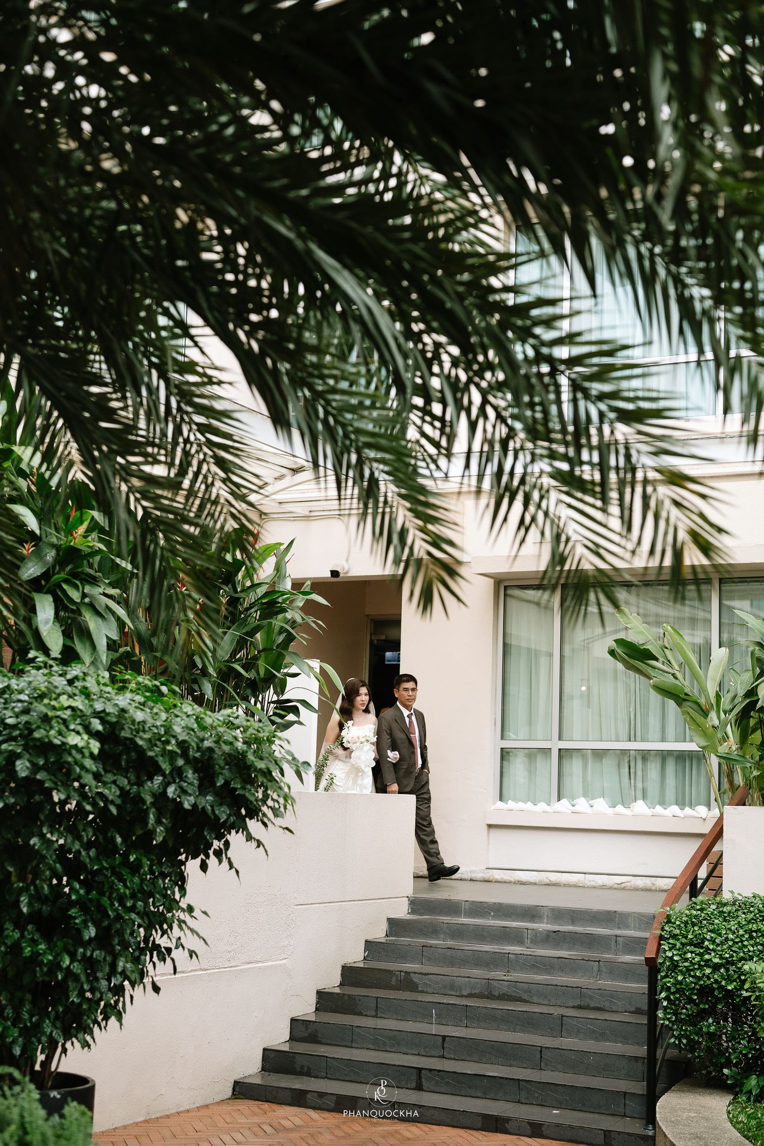 Bride in white gown and groom descend stone steps surrounded by lush tropical greenery at wedding venue.