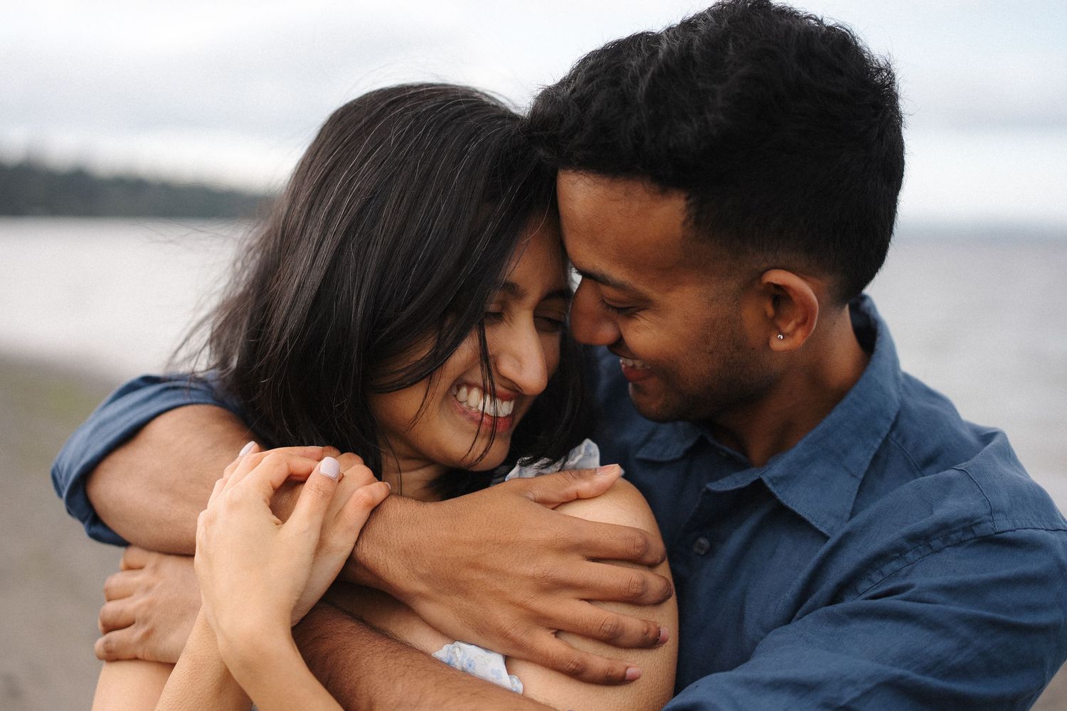 A romantic couple embrace and smile intimately together on a beach at sunset as waves roll in behind them.