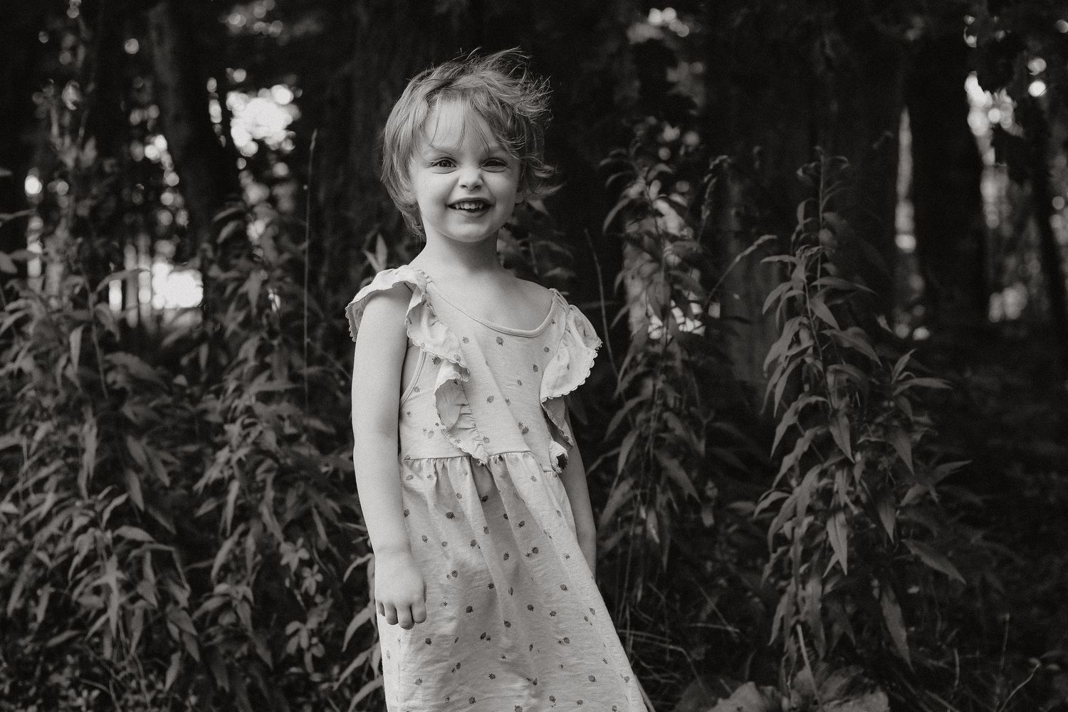 Young child in a delicate summer dress explores a natural outdoor setting among trees.