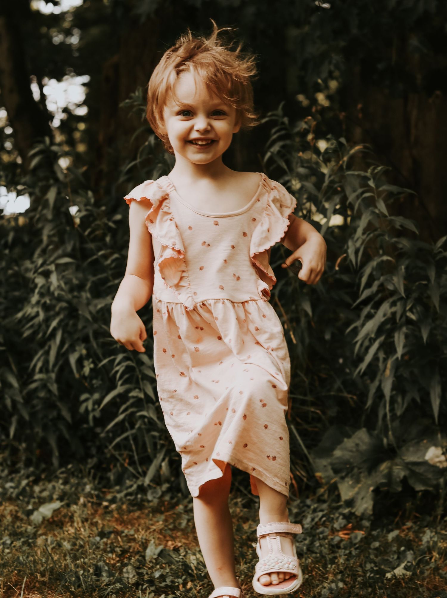 Small child in a ruffled pink dress twirls playfully in an outdoor wooded setting.