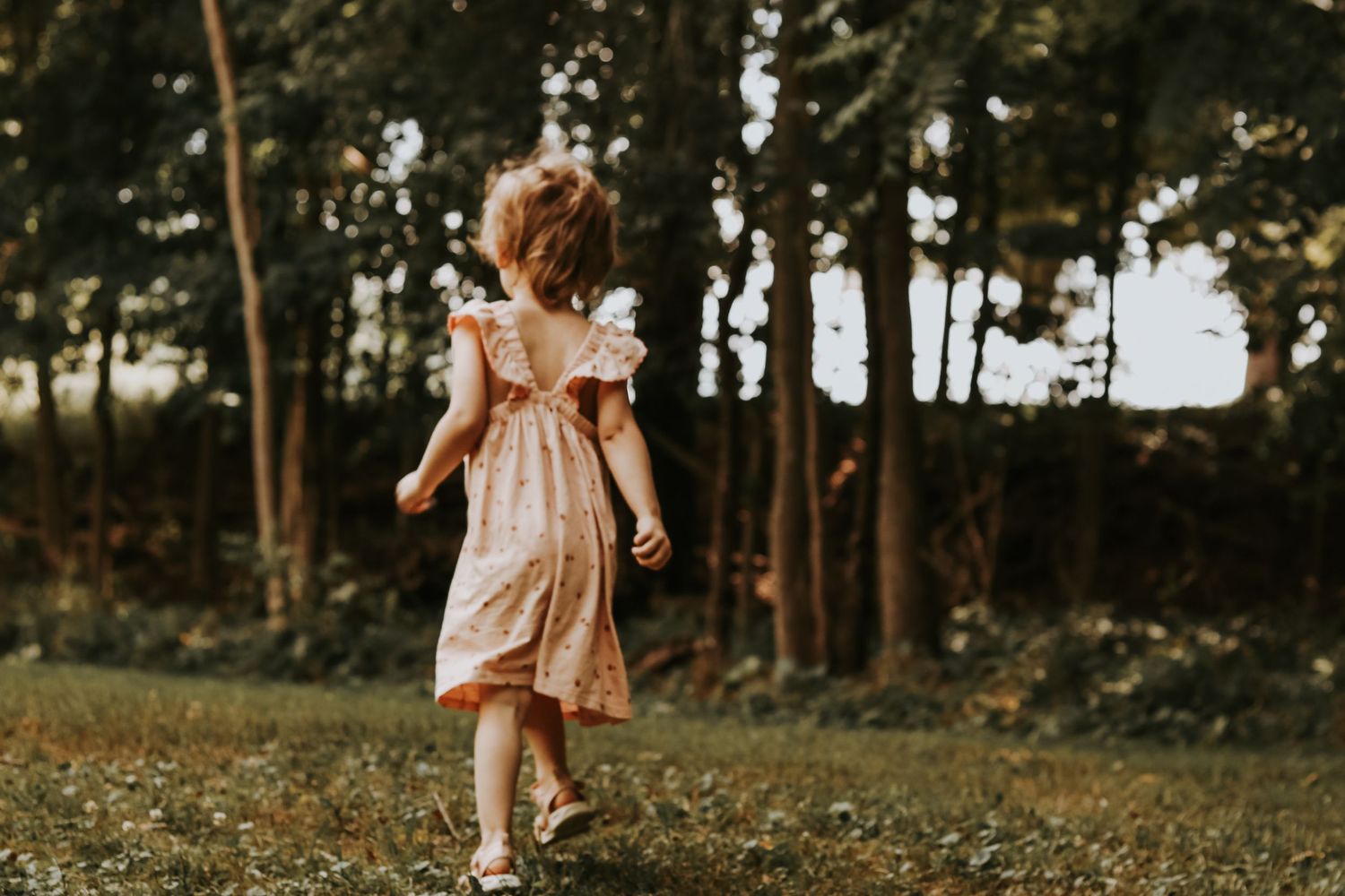 Child in a vintage-style dress dances through a grassy area with trees in the background.