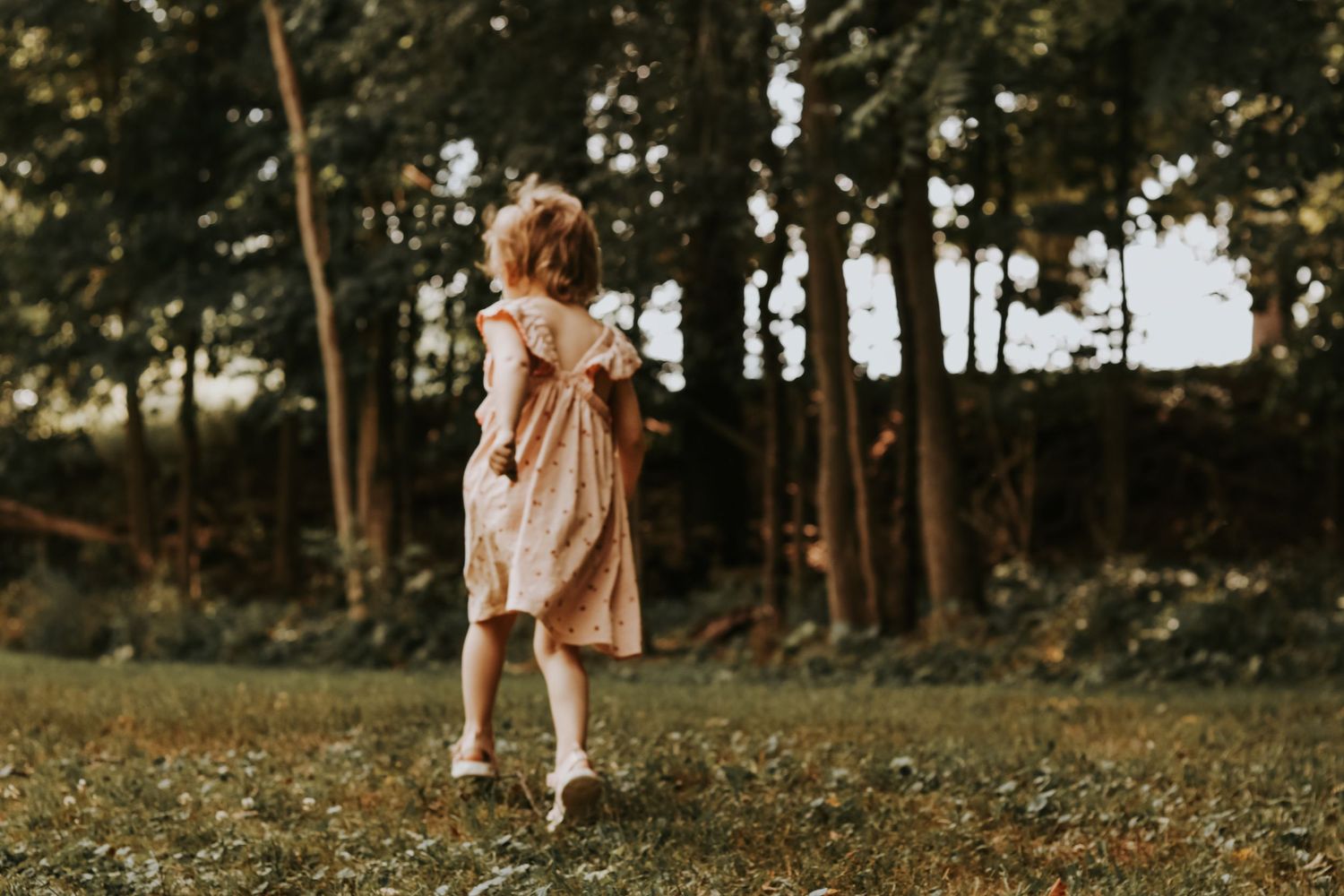Little one in a flowing dress plays freely in a wooded outdoor setting at sunset.