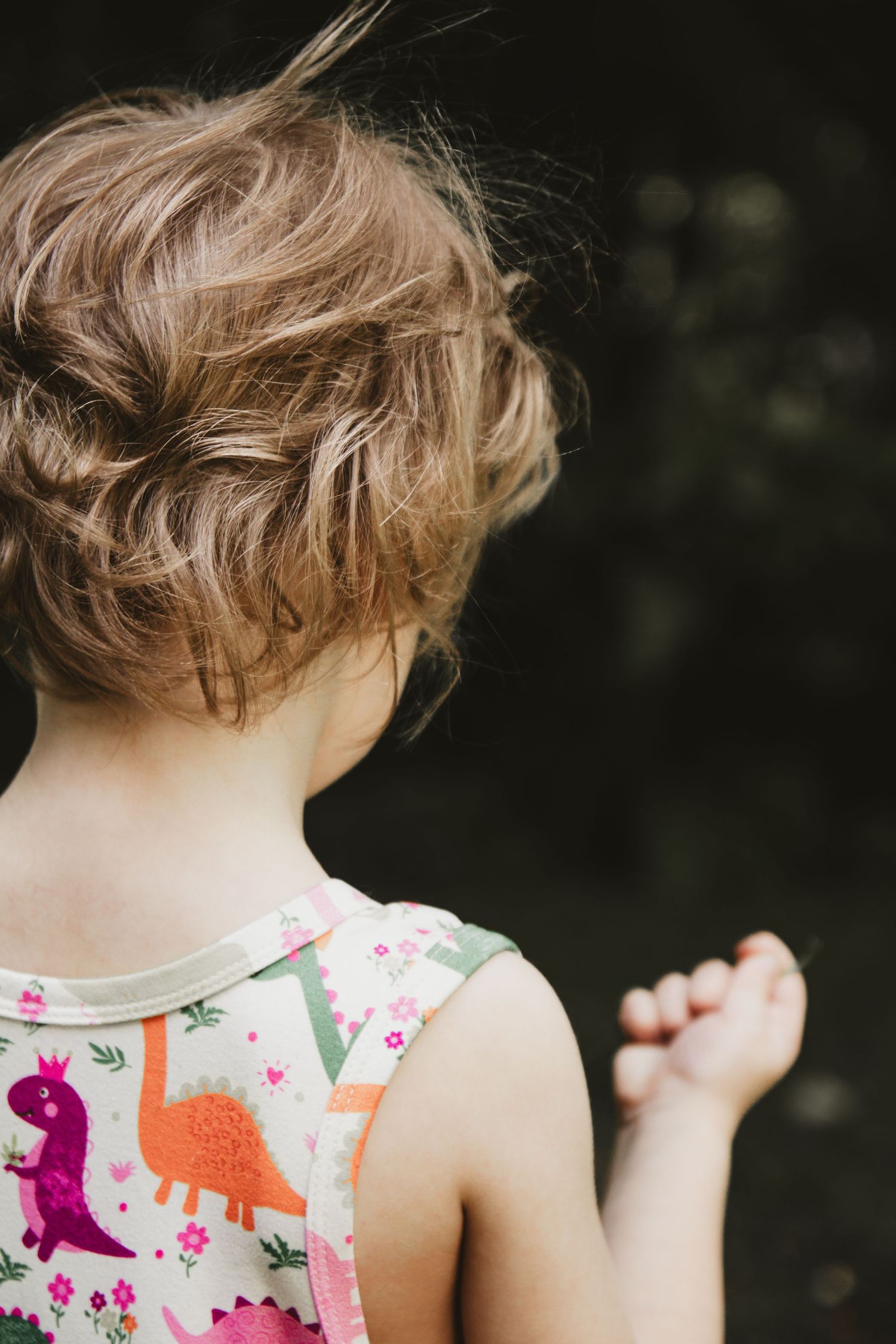 Back view of a child wearing a colorful dinosaur print sleeveless top against a dark background.