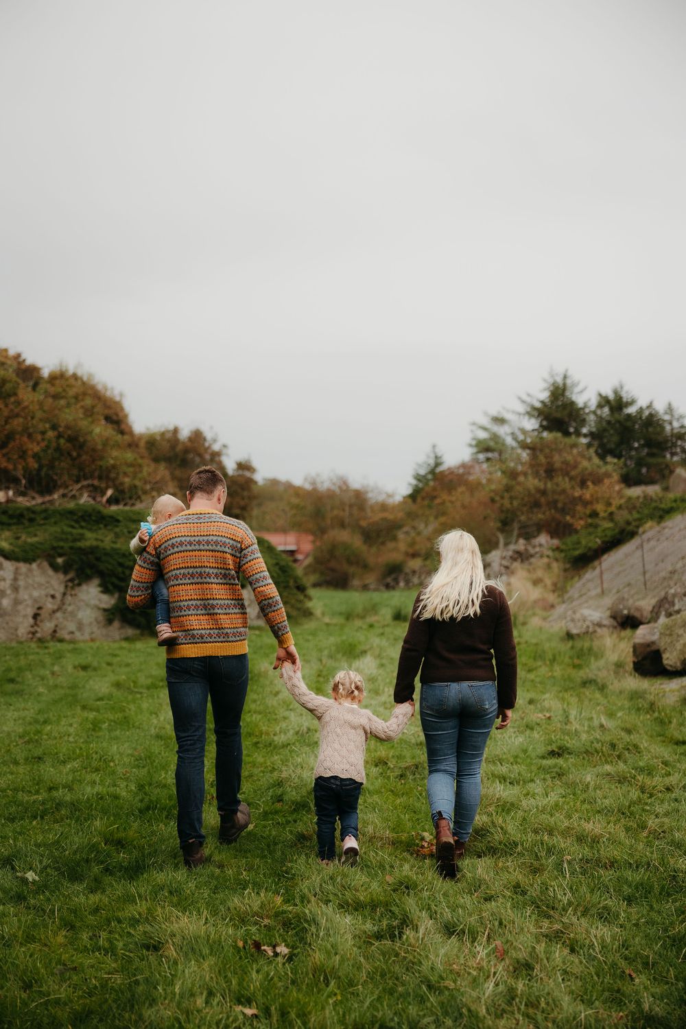 A family walks away across a grassy field holding hands, with a toddler in the middle and a baby carried by the father.
