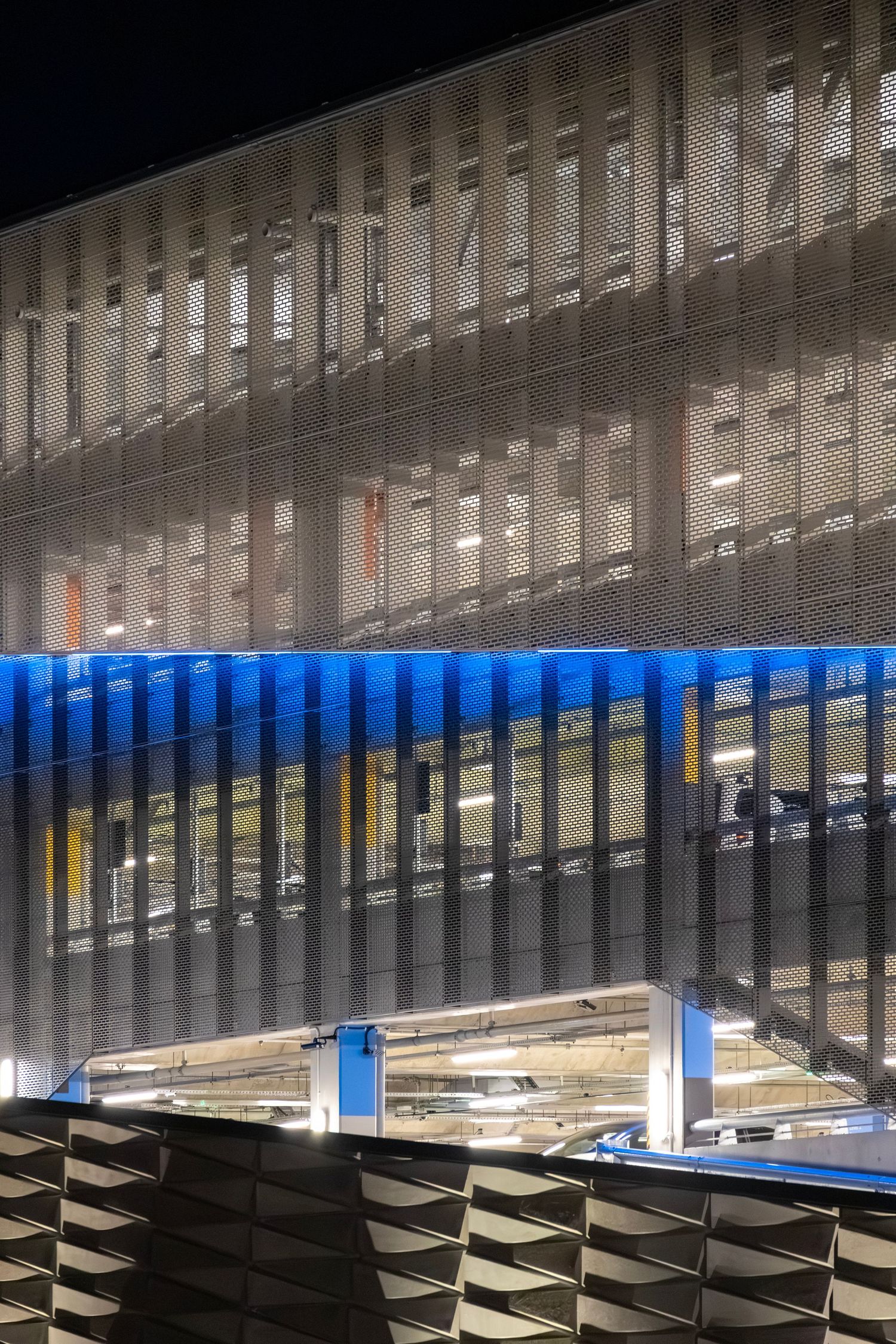 Multi-level car park at night with glowing interior lights and a blue-lit perforated metal screen facade.
