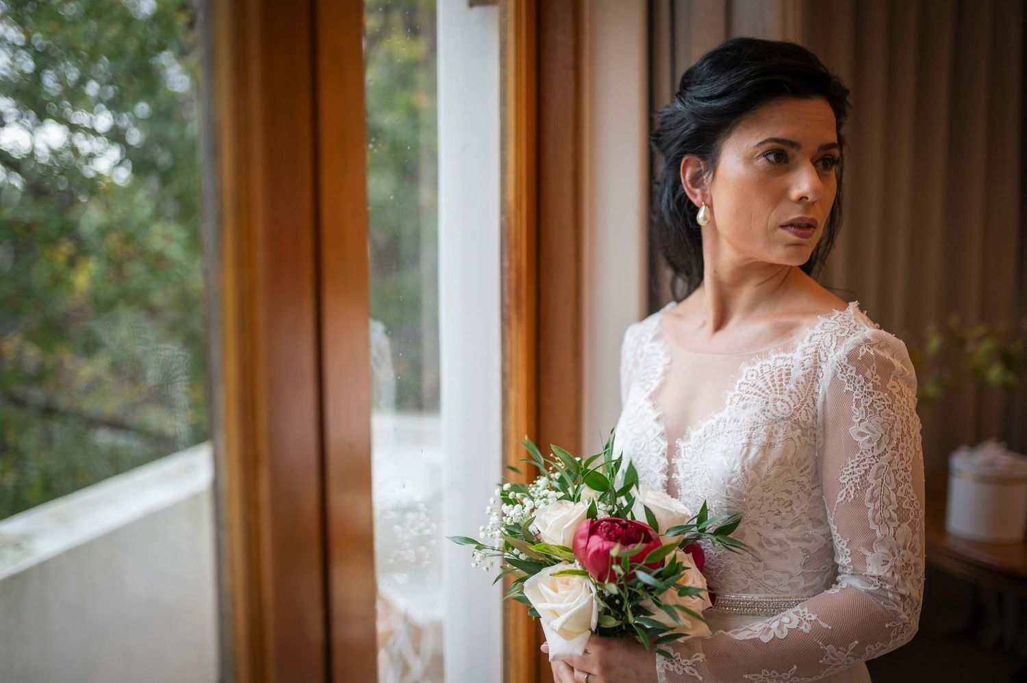 Beautiful bride in long sleeve lace wedding dress holds delicate bouquet while standing near window.