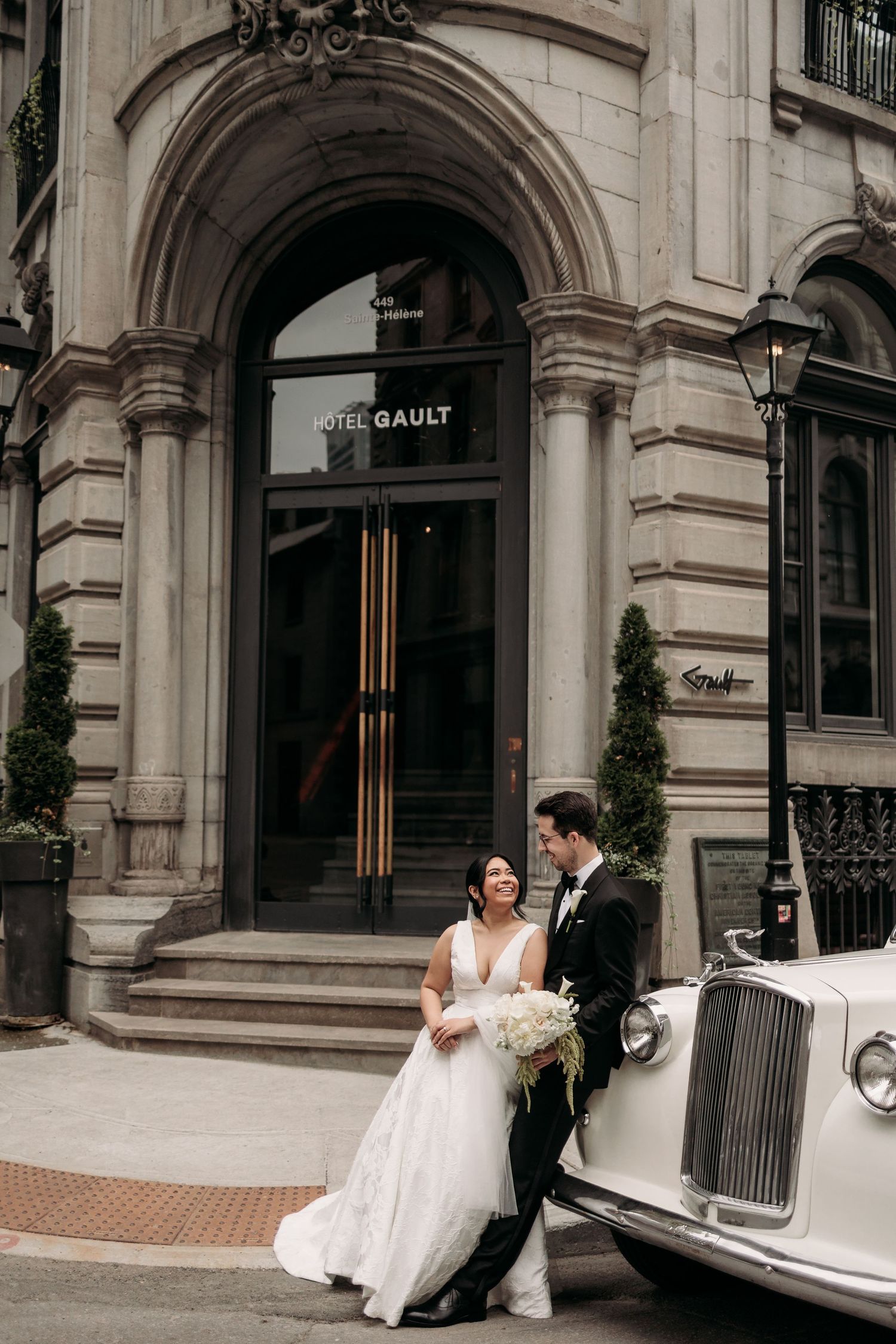 Bride and groom pose by a vintage white car outside Hotel Gault in elegant wedding attire. Captured by wedding photographer, Aliza Dworkind.