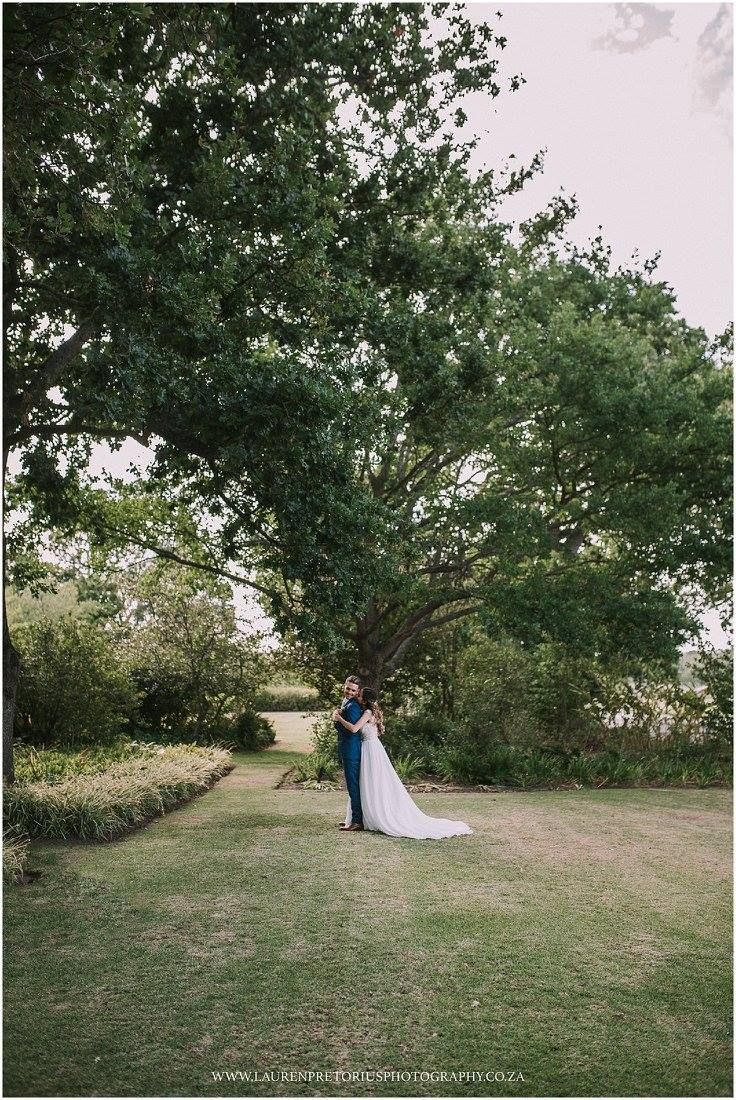 Newlyweds share an intimate moment under a large tree in a manicured garden setting.