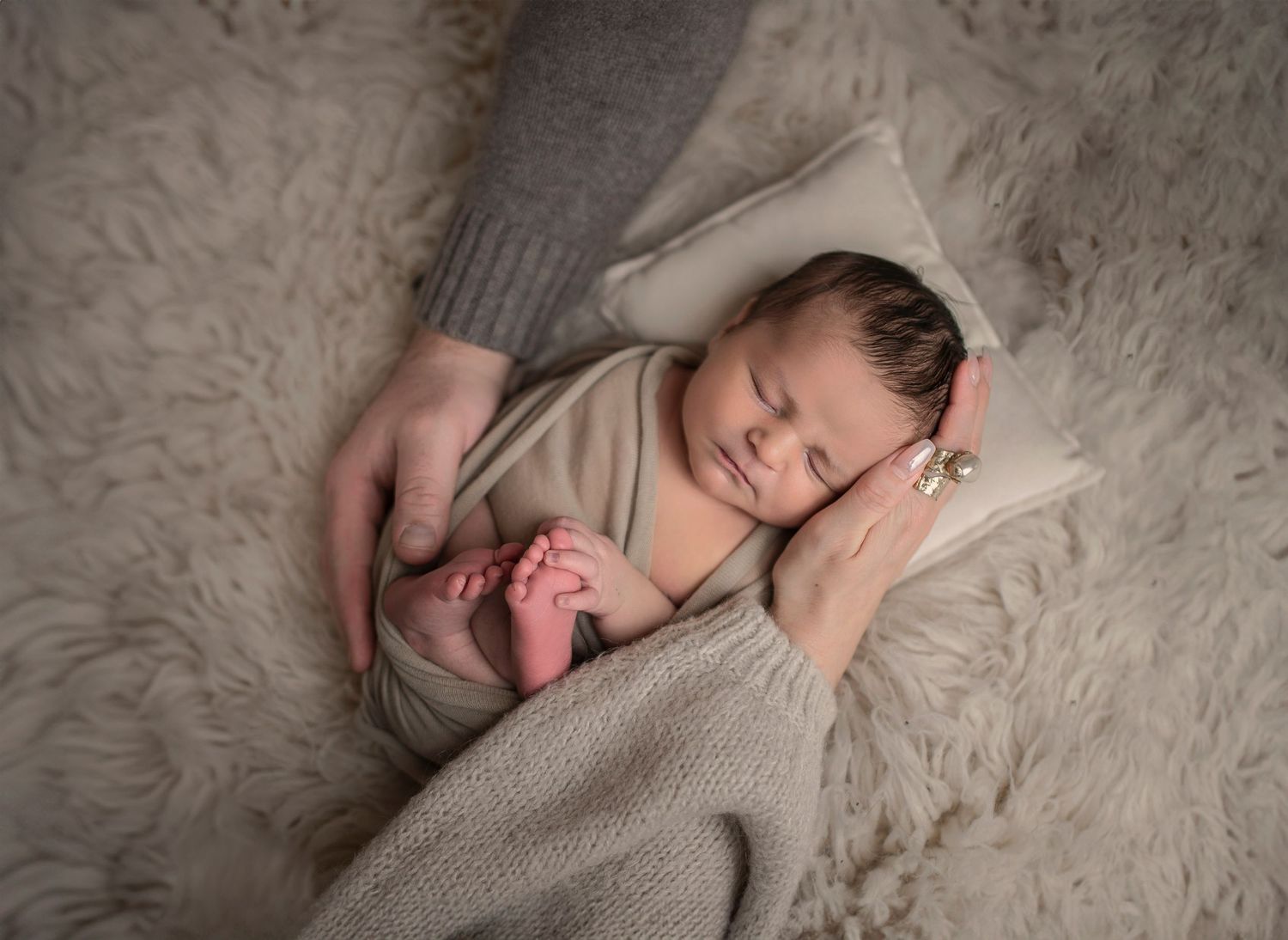 Sleeping newborn baby wrapped in beige swaddle blanket lying on soft white fur backdrop during professional photo session.