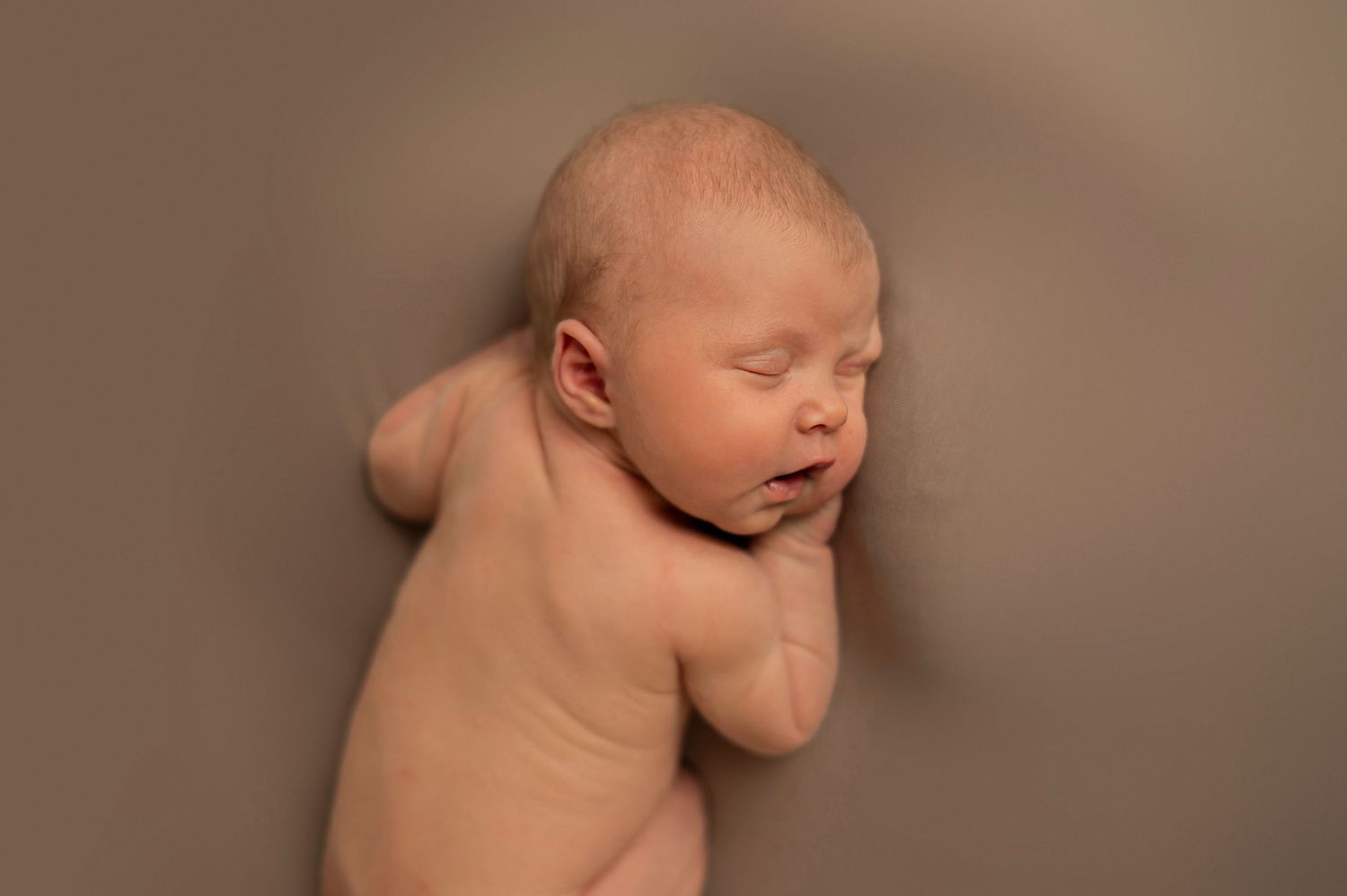 A sleeping newborn baby lies on soft brown fabric in a minimalist studio photography setup.