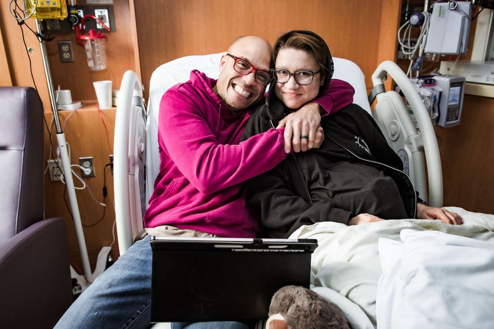 Hospital room scene with people sitting on chairs and medical equipment visible.