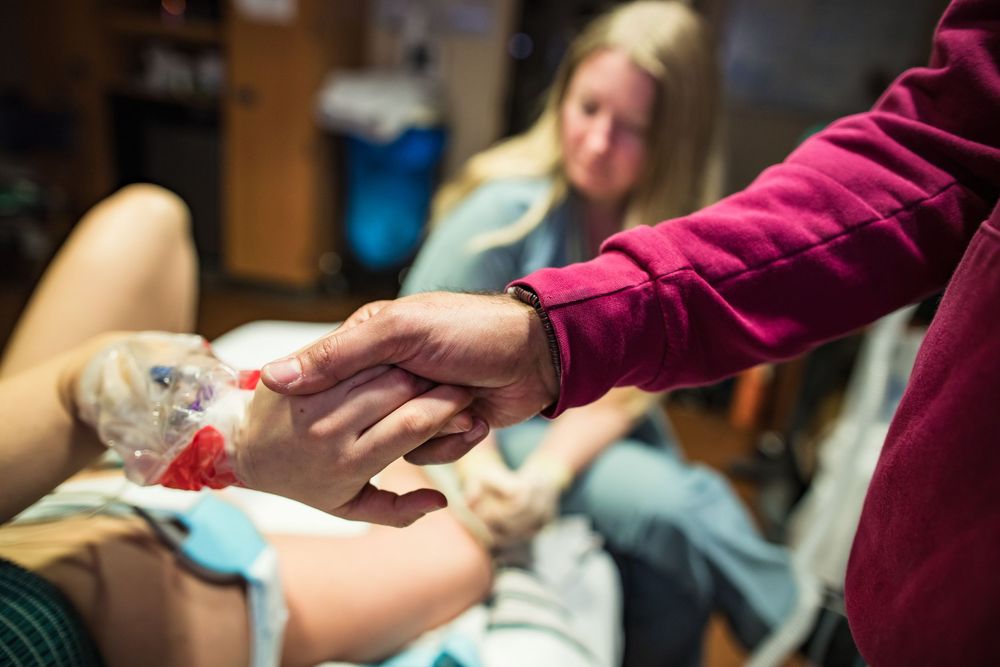 Healthcare worker in burgundy scrubs adjusting medical equipment at bedside.